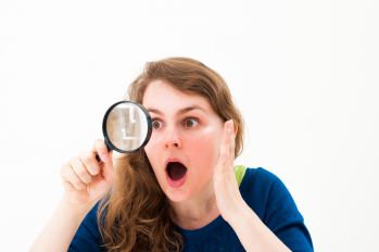 A woman with an expression of surprise and curiosity looking through a magnifying glass, symbolising the act of searching or investigating for something hidden or unclear.